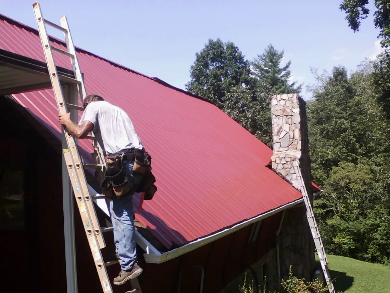 Roofer on a ladder beside a red metal roof and stone chimney — Soffit & Fascia Repair by Roofing Brothers in Cream Ridge